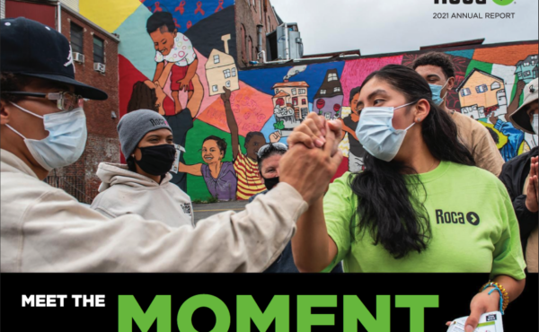 In the foreground, two people join hands, holding them up like a high five. In the background a mural of colorful and diverse portraits.